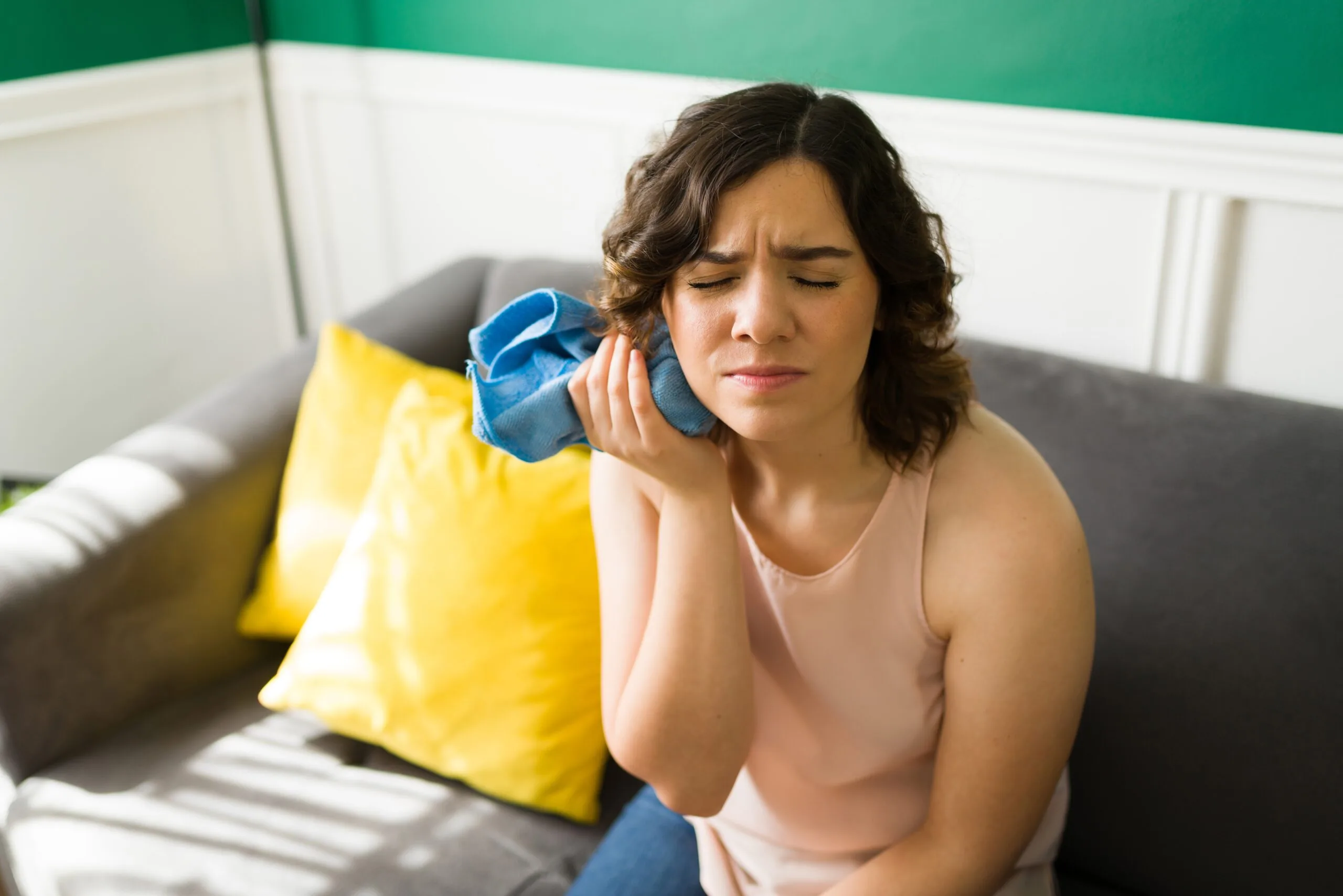 young woman sitting on a couch with a blue ice pack on her jaw, severe tooth pain